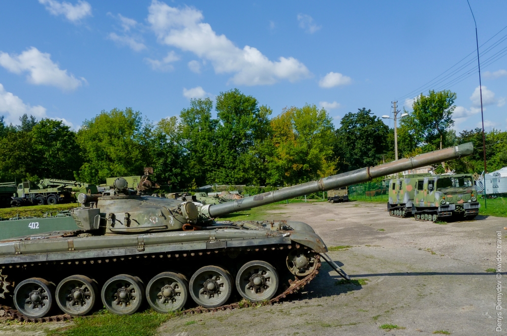 Фотография старого советского танка Т-72-М1, стоящего неподвижно на площадке в музее Каунаса среди зелёных деревьев и другой военной техники.