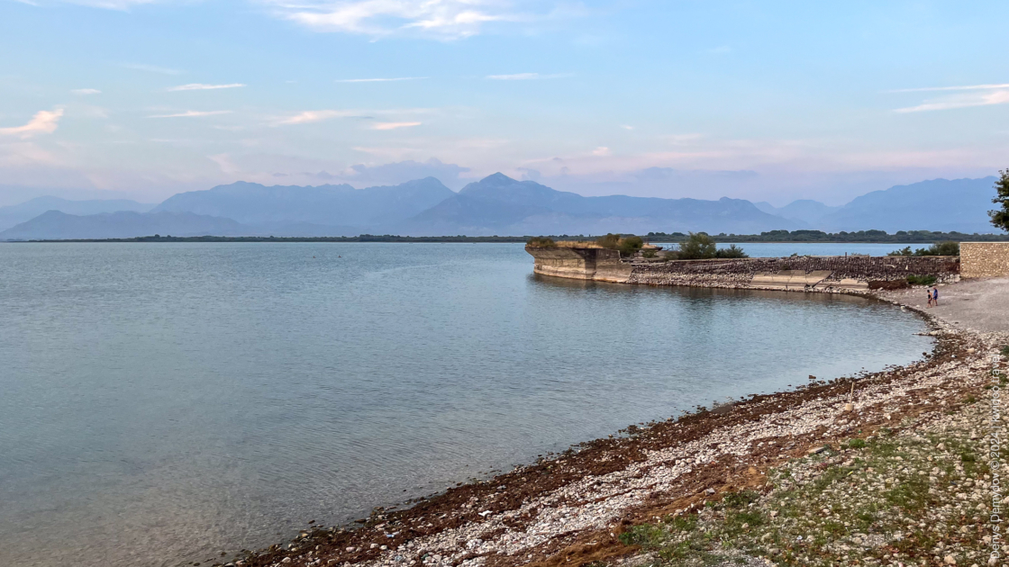 View of Skadar Lake and the mountains beyond. From the shore, an abandoned concrete garden is overgrown with weeds. 